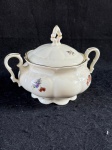 Hackefors. Açucareiro da marca Hackefors, uma empresa sueca de porcelana fundada em 1926. O açucareiro é feito de porcelana branca com detalhes em dourado e decorações florais. A peça possui duas alças laterais e uma tampa com um pequeno puxador. A base do açucareiro é ondulada, e o conjunto apresenta um design clássico e elegante, típico das peças da Hackefors.Conforme nas fotos.  