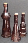 Três garrafas em vidro colorido prensado e trabalhado em relevo; podem ser usadas como decorativas ou para galheteiro. Peças na cor roxa ; medindo a maior 17 cm com a rolha e a menor 12 cm.