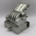COLECIONISMO - Miniatura de relógio em formato de antiga caixa registradora na cor prata em metal. Funcionando, porém sem garantias futuras. Medindo 6 x 5 x 7 cm.