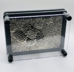 Caixa em acrílico com múltiplos pinos (alfinetes cegos) em metal que pressionado contra eles permite criar imagens e formatos. Ícone dos anos 80. Material: Metal e Acrílico. Medidas:  Altura 18,30 cm. Largura: 13,20 cm. Profundidade: 6,30 cm. Peso: 452 Gramas.