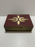 Caixa de madeira retangular, revestida de metal dourado e tampa decorada com figura estilizada sobre fundo vermelho. Interior revestido com tecido preto. Sinais de desgaste. Dimensões: 8 cm x 18 cm x 25 cm.