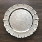 Belíssima bandeja em metal cinzelado decorada com flor de lis e estilizados vegetalistas incrustados em toda superficie e borda serrilhada. Dimensões: 44 cm.