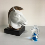 Duas estatuetas "Cabeça de cavalo", uma em porcelana art deco, de origem alemã e a outra em vidro (a escultura de porcelana está com desgaste e com restauro na crina). Dimensões: 18 cm (maior peça).