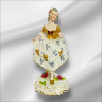 VIEIRA DE CASTRO - Escultura em porcelana VIEIRA DE CASTRO representando figura de dama antiga com vestes em policromia e vibrante ouro, apoiada sobre base em volutas com apliques florais. Possui registro. Mede 26cm altura.