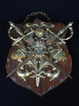 Escudo Medieval decorativo de madeira com "espadas" entrecruzadas, lâminas de ferro sendo apoiadas por aguia de duas cabeças, sobre elmo da armadura e dois guerreiros montados em cavalos. Espadas com marcas do tempo e águia sem uma das asas. 25x18 cm
