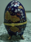 Caixa porta joia de porcelana colorida na forma de ovo, decorada com flores e personagens, guarnição de metal. Alt. 9,5cm