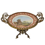 Fruteira com prato circular em porcelana policromada, ornado por cena campestre bordada por faixa floral sobre fundo coral. Montagem em bronze ormolu com borda em godrões, ladeada por alças duplas reviradas em arabescos e realçadas por cachos de uva. Base com suporte circular, centralizando colunata canelada apoiada em pernas inclinadas, recamadas por folhagens e rematadas por pés em volutas. 20 x 31 x 25 cm.