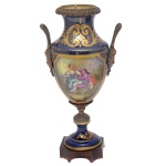 Vaso em porcelana europeia policromada, de formato cônico, com montagem em bronze, ornado em reservas por cena galante à frente e paisagem no verso. Fundo azul Bleu du Roi, realçado por volutas e adornos dourados. Bojo abaulado com pegas laterais, encimado por gargalo retraído em corneta e borda revirada. Haste anelada apoiada em base circular, sobre peanha quadrangular com quinas arqueadas. 28 x 15 cm.