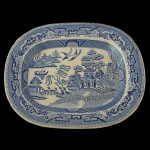 Travessa em louça inglesa de formato oblongo e borda lisa. Apresenta decoração em esmalte de tom azul sobre fundo branco no padrão conhecido como Pombinhos. Ao centro cena lacustre com casario e arvoredo. 36 x 29 cm.
