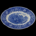 ARABIA - Travessa oval em louça inglesa, decorada em azul. Apresenta borda ondulada com fino friso estilizado e margem ornada por flores e folhagens. Centro apresentando cena rural com rio e montanhas, castelo e edificações, e figuras com gado. Marca da manufatura no verso. 36 x 25 cm. Bicado na borda.