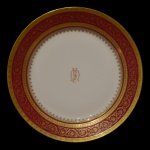 LIMOGES, UNIC  Travessa circular em porcelana francesa branca, com borda circular ornamentada em esmaltes vinhos e dourados. Apresenta frisos geométricos e faixa com volutas. Centro realçado por monograma. No verso marca da manufatura e da Casa12, Rue de Paradis, Paris. 33 cm.