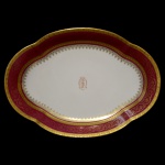 LIMOGES, UNIC - Travessa de formato oblongo em porcelana francesa branca, com borda circular ornamentada em esmaltes vinhos e dourados. Apresenta frisos geométricos e faixa com volutas. Centro realçado por monograma. No verso marca da manufatura e da Casa12, Rue de Paradis, Paris. 41 x 29 cm.