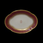 LIMOGES, UNIC - Travessa de formato oblongo em porcelana francesa branca, com borda circular ornamentada em esmaltes vinhos e dourados. Apresenta frisos geométricos e faixa com volutas. Centro realçado por monograma. No verso marca da manufatura e da Casa12, Rue de Paradis, Paris.. 36 x 26 cm.