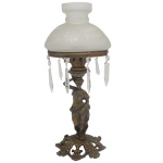 Luminária de mesa em bronze polido. Haste no formato de figura feminina sustentando sobre a cabeça vaso com largo coroa saliente, ornada à volta por pingentes em cristal. Cúpula em vidro leitoso, encimada por pequeno gargalo com borda alta e reta. Base circular, lavrada e vazada com adornos vegetais. 48 x 17 cm.