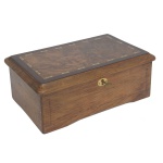 Caixa de música em madeira lisa, de formato retangular. Tampa com borda saliente, ornada por friso geométrico em marqueterie. Interior com rolo sob vidro, ladeado por alavanca de corda e botões seletores. 12 x 30 x 18 cm.