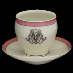 Xícara com seu pires em porcelana. Superficie de cor branca com friso rosa e iniciais entrelaçadas. 6 x 6 cm (xícara). 11 cm (pires).
