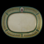 Travessa ovalada com laterais retas, em porcelana branca. Apresenta larga margem decorada em várias tonalidades verdes e ouro. Frisos geométricos bordando faixa com arcos entrecruzados, interrompida por medalhões, sendo o do topo realçado por brasão monogramado. Fio de cabelo e restauro. 32 x 25 cm.
