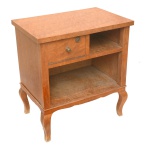 Mesa de cabeceira retangular, em rádica clara, com tampo liso apoiado em caixa reta. Frente com compartimento aberto, encimado por gaveta com chave ladeada por pequeno nicho. Pernas curvilíneas com joelhos salientes.