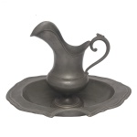 Conjunto de barbear em estanho liso, realçado por filetes canelados e frisos em godrões. Constando de jarra de formato balaústre com largo bico recortado e revirado. Alça lateral em S e base circular. Acompanha prato oval fundo, com borda ondulada e característico recorte à frente. 6 x 38 x 30 cm. B) Jarra para água.  Alças curvas. 25 x 23 cm. Sinais de uso e do tempo.