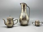 METAL  Conjunto com bule, açucareiro e jarra alta - Conjunto composto por três peças antigas em metal espessurado a prata: Bule com tampa fixa, bico decorado, alça curva e quatro pés em forma de concha  desenho clássico com elegantes relevos rococó. - Açucareiro com tampa, duas alças e pés ornamentados, formando par com o bule. - Jarra alta, de corpo bojudo e pescoço longo, com alça reta e base estriada. Design limpo e robusto, com pátina do tempo. Med. maior 26 cm e menor 7 cm. Desgastes.