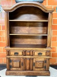 MOBILIÁRIO - CRISTALEIRA / ESTANTE RÚSTICA EM MADEIRA MACIÇACharmoso móvel em madeira maciça com acabamento rústico, composto por parte superior com prateleiras abertas e moldura superior arqueada, ideal para livros, objetos decorativos ou utensílios. A parte inferior possui duas gavetas com puxadores em metal envelhecido e duas portas com ferragens em ferro forjado, revelando bom espaço interno para armazenamento. Design inspirado no estilo colonial, com rica presença de molduras e entalhes em relevo. Excelente peça para ambientes com decoração clássica, rústica ou de fazenda.  Med. 213x140x49 cm.