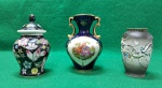Lote contendo 3 pequenas peças em porcelana, sendo; à esquerda, miniatura de pote japonês, na cor  preta com tampa, decorado com flores coloridas e borboletas; ao  centro, miniatura de ânfora na cor azul cobalto, com alças douradas. e decorada ao centro com cena galante;  à direita, miniatura de pote chinêso, na cor cinza e em  relevo,  desenhos intrincados de um dragão. Usadoa. Todos em ótimo estado.