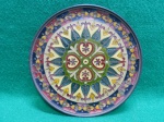 Prato decorativo em bronze, apresenta um padrão central em forma de trevo com corações, cercado por elementos florais e folhagens coloridas, predominando tons de verde, amarelo, vermelho e azul, com detalhes em dourado. Usado, em muito bom estado. Mede 16,5 cm diâmetro.