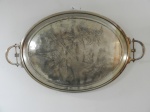 Grande bandeja em metal prateado, decorada ao centro com gravações de plantas e árvores. Seu centro não se encontra totalmente plano, apresentando pequenos amassados. mede 80 C x 50 L.