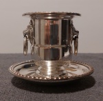 Pequeno pote sobre sua bandeja, em metal banhado a prata, decorado em suas laterais com cabeça de leão, transpassada por argolas. A peça tem medida total de 8 A x 9,5 diâmetro. Usado. Em ótimo estado.