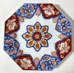 CIA DAS ÍNDIAS- IMARI-JAPÃO SÉCULO XIX - Prato de formato octogonal, em porcelana policromada no estilo Imari, decorado em vermelho-ferrugem, azul cobalto e dourado. No centro, composição floral em forma de roseta, cercada por reservas com motivos florais e estilizações orientais. A paleta e a simetria ornamental revelam a elegância típica da produção japonesa destinada à exportação no período Edo/Meiji. Medidas: