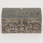 METAL PRATEADO, uma (1) caixa porta joias com intrincada ornamentação relevada em elementos vegetalistas, formato de bau, interior com forração aveludada na tonalidade vermelha, medindo 10,5 x 6,5 x 5,5 cm, vide foto.