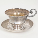 METAL PRATEADO, uma (1) xícara com pires, ornamentação em baixo relevo em elementos vegetalistas, alça relevada em folhas e volutas, manufatura EBERLE (Abramo Eberle 1880, Monte Magrè 1945, Caxias do Sul, veio com uma das primeiras levas de imigrantes italianos para o Brasil em 1884 junto com seus pais Giuzeppe Eberle e Luigia Zanrosso, seu pai abriu uma pequena funilaria mas como tinha que trabalhar na lavoura quem cuidava era a mãe e o pequeno Abramo ajudava na funilaria; em 1896 quando o pai resolveu vender a funilaria para concentrar os esforços da família na lavoura Abramo, com 16 anos de idade, se propôs a comprá-la do pai, em 1918 já com o nome Abramo Eberle & Cia iniciou a fabricação de talheres e utensílios de mesa, morreu em 1945 com 65 anos de idade e os filhos deram continuidade aos negócios até 1984 quando venderam para o Grupo Zivi), pires com mossas, sinais de uso, vide foto.