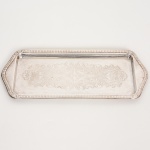 METAL PRATEADO, uma (1) bandeja quase retangular, confeccionada em aço inox na técnica de estamparia, borda relevada em corda, conchas, volutas e elementos vegetalistas, fundo com ornamentação em baixo relevo, medindo 33,5 x 14 cm, sinais de uso, vide foto.