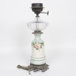 Candeeiro de mesa com base em metal fundido com ornamentos em relevo, corpo central em porcelana esmaltada decorada com pintura floral e reservatório superior em vidro transparente moldado em formato abobadado. Peça originalmente a querosene, revertida para eletricidade. Apresenta oxidação nas partes metálicas e fiação original sem teste funcional. Med.: Alt. 49 x Diam. 16 cm.