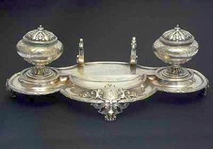 Domitila de Castro Canto e Mello, Marquesa de Santos. Raríssimo <b>tinteiro de bandeja</b>  recortada em prata inglesa vitoriana com as armas da marquesa de Santos. Apresenta dois recipientes laterais em forma de urnas, gravadas com estilizações e bordas filetadas intercaladas por "X". Parte posterior com suporte para pena. Um dos suportes faltando parte e um dos recipientes faltando o vidro azul. Contraste da cidade de Londres de 1845 e marca do prateiro real ROBERT GARRARD. Compr. 22 cm