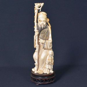 <b>Escultura japonesa </b>em marfim, representando ancião com bastão e criança ao lado. Figura masculina idosa de expressão serena e sábia, trajando vestes tradicionais minuciosamente entalhadas. Sustenta bastão decorado por dragão na extremidade superior e segura leque na outra mão. Ao seu lado, criança em pose respeitosa. Apresenta marca na parte posterior. Alt. 27 cm