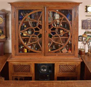 <b>Móvel bar vitrine </b>em madeira entalhada, de grandes proporções, composto por parte superior com portas envidraçadas adornadas por elaborada grade circular geométrica. A base apresenta compartimentos laterais fechados por treliças de madeira, além de nichos cilíndricos para armazenamento de garrafas e espaço central aberto.  Med. 240 x 162 x 43 cm