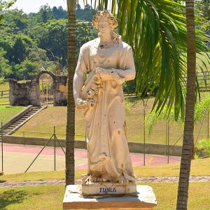 <b>Escultura </b>em faiança representando figura feminina alegórica intitulada "Flora", em posição ereta, portando grinalda de flores ao braço e vestes esvoaçantes, apoiada sobre base retangular. Inscrição "José P. Valente / Fª S?? Antônio  Porto". Apresenta desgastes e lascados pontuais compatíveis com exposição externa.