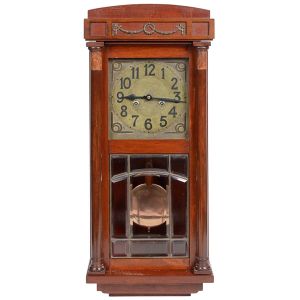 <b>Relógio de parede </b>em madeira nobre, com mostrador metálico e mecanismo de pêndulo visível através de porta envidraçada com delicados recortes em vidro bisotado. Estrutura adornada com delicados frisos e colunas laterais, encimada por frontão suavemente arqueado com aplicação metálica decorativa. Med. 78 x 33 cm