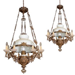 <b>Par de lustres </b>em metal dourado ricamente trabalhado, com corpo central vazado em motivos fitomorfos e reservas com querubins, sustentando aberturas para vela eletrificada com "bobèches" em vidro fosco. Estrutura superior com corrente dupla decorada, sustentando cúpula em vidro leitoso. Alt. 120 cm