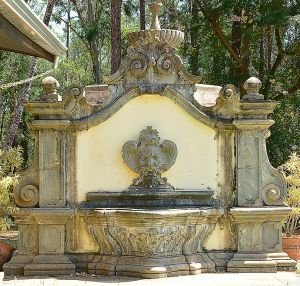<b>Fonte monumental </b>em pedra lavrada, de inspiração barroca, composta por grande painel recortado com volutas laterais e coroamento em bacia superior. Ao centro, carranca em alto-relevo verte água para ampla bacia sinuosa inferior, ricamente ornada com motivos fitomórficos. Compr. total 396 cm