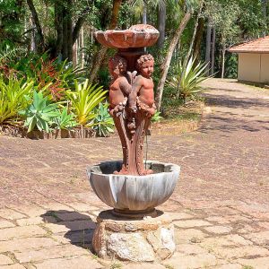 <b>Chafariz </b>em ferro fundido, composto por bacia inferior em volutas apoiada sobre base em pedra e fuste ornado por quatro figuras de "putti" em relevo, interligadas por guirlandas de frutos. A parte superior apresenta reservatório circular em forma de concha, também em pedra. Alt. 147 cm