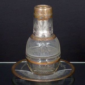 <b>Conjunto de moringa com copo e prato </b>em cristal lapidado, com guarnições em prata francesa, com bordas e arremates cinzelados em friso floral. Corpo da garrafa e do copo decorados com gravações em motivos fitomórficos. O prato apresenta fissura visível. Contraste "Cabeça de Minerva". Alt. 22 cm