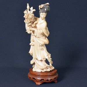 <b>Escultura em marfim </b>representando gueixa, adornada com trajes longos e esvoaçantes. O penteado elaborado da figura é cuidadosamente esculpido, e suas duas mãos seguram uma flor de peônia, que simboliza riqueza, boa sorte e florescimento. Base em madeira entalhada. Alt. 27 cm