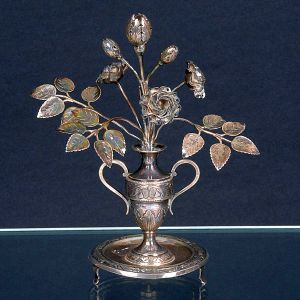 <b>Paliteiro </b>em prata portuguesa, modelado em forma de arranjo de rosas com folhas delicadamente cinzeladas, inseridas em vaso de duas alças decorado com frisos e volutas. Base circular apoiada sobre pés em voluta. Contraste da cidade do Porto e marca de prateiro. Alt. 21 cm