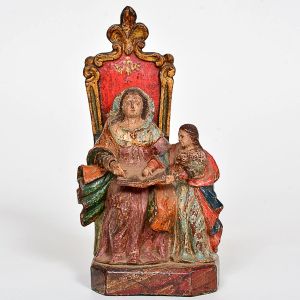 Imagem de <B>Sant'Ana Mestra </B>em madeira entalhada e policromada, representada sentada em trono de espaldar alto em vermelho e dourado, de perfil recortado. A Santa segura livro aberto sobre o colo enquanto orienta a pequena Virgem Maria, posicionada ao seu lado. Alt. 18 cm