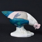 <b>Escultura em porcelana </b>europeia Art Déco, representando galinha de capoeira em posição de forrageio, esmaltada em tons de azul-petróleo, cinza, rosa e branco. Base marcada na parte inferior com carimbo coroado e iniciais "M & C", além de numeração em relevo. Alt. 8 cm