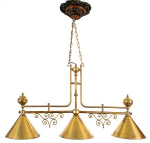<b>Luminária pendente tripla</b>, para mesa de bilhar, com estrutura em latão, braços ornados em volutas e cúpulas cônicas decoradas internamente em esmalte verde. Suspensa por correntes ligadas a canopla em madeira entalhada com pintura patinada. Med. 65 x 105 cm