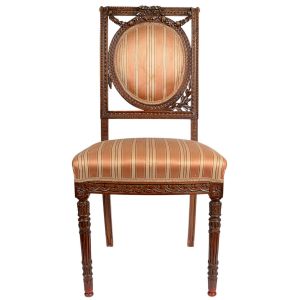 <b>Cadeira </b>em madeira entalhada, com espaldar emoldurado por motivos florais, encimado por grinalda de laços e flores. Assento estofado em tecido listrado em tons de dourado e pêssego. Estilo Luís XVI. Apresenta perda em parte do entalhe. Alt. 93 cm