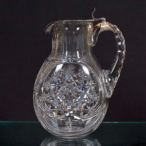 <b>Jarra </b>em cristal com tampa articulada em metal. Corpo bojudo ricamente lapidado em padrões geométricos e florais em alto-relevo, com alça facetada e bico com acabamento superior em metal. Monograma gravado e datado "25-4-1921".